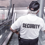 A man wearing a white shirt with black lettering that says security on the back going down an escalator.