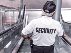 A man wearing a white shirt with black lettering that says security on the back going down an escalator.