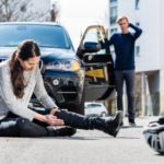 A woman on the ground in pain holding her leg next to a bicycle and a helmet. Man in the foreground stressed standing by his car.