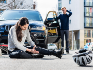 A woman on the ground in pain holding her leg next to a bicycle and a helmet. Man in the foreground stressed standing by his car.